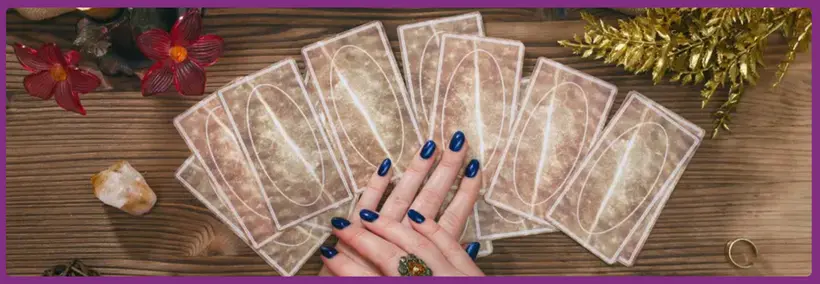Hands with dark blue nail polish positioned over a spread of tarot cards arranged in a fan pattern on a wooden surface, surrounded by decorative elements including red flowers, crystals, and golden tinsel.