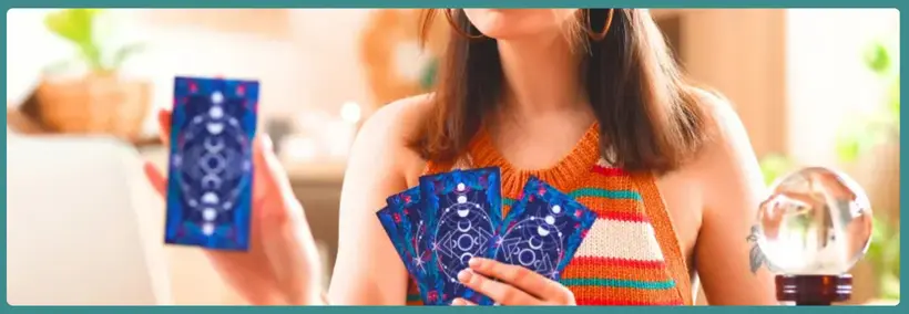 Woman wearing a colorful striped top holding blue tarot cards in both hands, with a crystal ball visible on the table beside her.