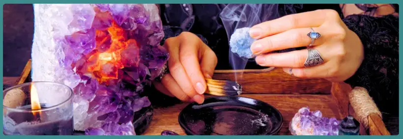 Hands performing a ritual or divination practice over a dark scrying bowl, surrounded by purple amethyst crystals, lit candles, and mystical items with smoke or incense visible.